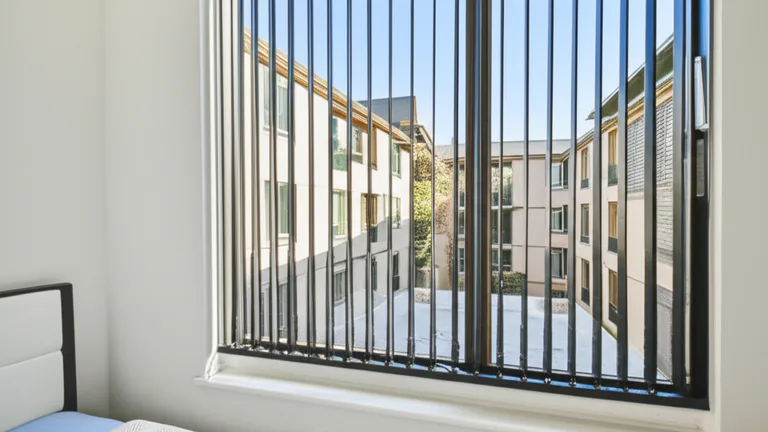 Egress-compliant telescopic steel window bar installed in multifamily apartment bedroom with quick-release mechanism visible, morning light, ground-floor unit